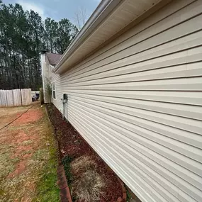 Side view of a beige vinyl-sided house with a landscaped garden bed featuring red mulch, bordered by bricks, adjacent to a patchy lawn and a wooden fence under cloudy skies.