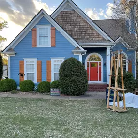 A charming blue house with orange shutters and a vibrant red front door stands proudly under a partly cloudy sky. The well-maintained lawn and green bushes enhance its curb appeal.