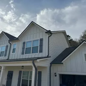 A two-story white farmhouse townhome with dark gray shutters and gutters is captured under a cloudy sky. The house features a prominent front-facing garage.