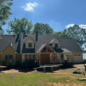 A waterfront house undergoing exterior renovation, with new roofing visible and sections still under construction. Construction materials are scattered around the yard, indicating active work.