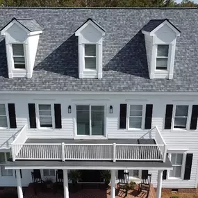 An aerial view of a large white colonial-style house with a new light gray shingle roof, featuring three prominent dormer windows. Black shutters provide a stark contrast.