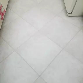 This image shows a clean, professionally restored tile floor by The Groutsmith. The large, light-colored tiles feature precise, uniform grout lines, giving the kitchen area a refreshed and polished appearance.