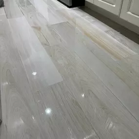 This image features a glossy floor of light-colored wood-look plank tiles, reflecting light and showing subtle wood grain patterns, extending towards white cabinets and a dark appliance. The impeccable shine suggests professional cleaning and sealing services by The Groutsmith.