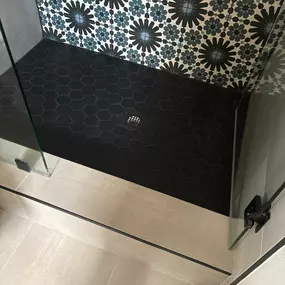 This image showcases a modern shower with a dark hexagonal tile floor and a striking patterned wall featuring black and blue floral designs. Clear glass shower doors frame the impeccably clean and vibrant tile work, highlighting professional tile and grout care.