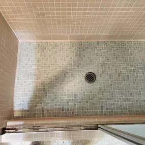 This image shows a shower with beige square wall tiles and a mosaic-patterned floor in shades of gray and white, with a central drain. Noticeable dirt and mildew in the grout lines highlight the need for professional cleaning and restoration by The Groutsmith.