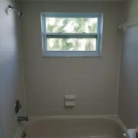 This image showcases a clean and well-maintained bathroom shower and tub area, featuring light-colored square tiles with pristine white grout. A window above the tub allows natural light, highlighting the spotless condition and recent professional care by The Groutsmith.
