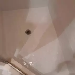 A clean, white cultured marble shower with a visible drain and a partial view of a glass door. The shower walls are tiled, reflecting light, suggesting a recently cleaned or well-maintained surface by The Groutsmith.