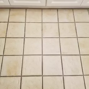 This image shows a close-up of light-colored floor tiles with dark, possibly discolored grout lines underneath white kitchen cabinets. It highlights a common problem area, demonstrating the need for professional grout cleaning and restoration services from The Groutsmith.