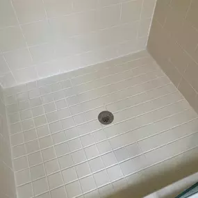 This image shows a perfectly clean and bright shower pan and walls, featuring light beige square tiles with impeccably white grout lines and a central drain. The pristine condition highlights the superior results achievable with professional tile and grout services from The Groutsmith.