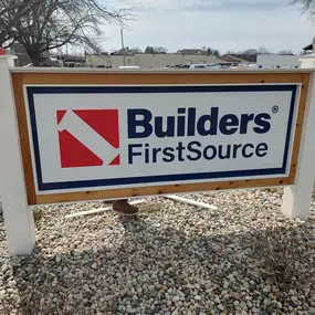 Close-up of a large Builders FirstSource outdoor sign mounted on white posts with a wooden frame. The sign sits on a bed of landscaping rocks near a grassy area, with part of a utility truck and a building visible in the background. The surrounding environment suggests a commercial or industrial area.