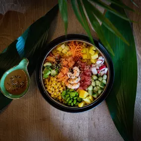 Frische Poké Bowl mit Garnelen, Mango, Edamame, Avocado, Rotkohl und Karotten, serviert mit einem Dip auf tropischen Blättern angerichtet.