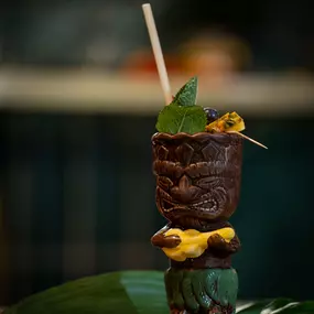 Exotischer Tiki-Cocktail im verzierten Becher mit Limette, Minze und Trinkhalm – stimmungsvoll angerichtet auf tropischem Blatt.