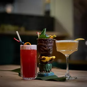 Drei stilvolle Cocktails in verschiedenen Gläsern: fruchtiger Longdrink, exotischer Tiki-Cocktail und eleganter Sour – serviert in modernem Bar-Ambiente.