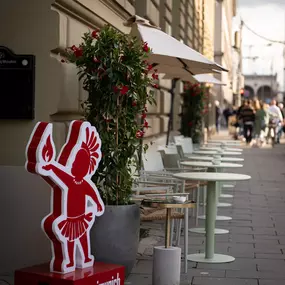 Einladender Außenbereich eines Restaurants mit Tischen, Sonnenschirmen und auffälliger roter Werbefigur – stilvolle Straßenszene mit mediterranem Flair.