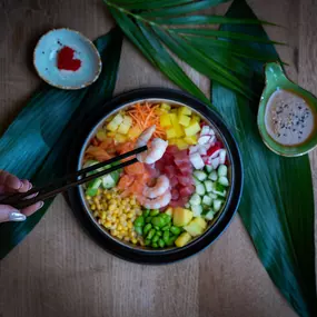 Bunte Poké Bowl mit Garnelen, Mais, Edamame, Mango und Karotten – liebevoll serviert auf tropischen Blättern mit Dips und Stäbchen.