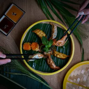 Asiatische Spezialitätenplatte mit Gyoza, Shrimps und Gemüse, serviert auf Bambusteller mit Dip und dekorativem Palmblatt – zwei Personen greifen mit Stäbchen zu.