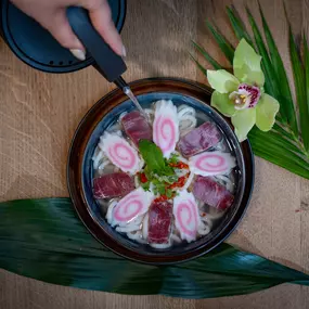 Kunstvoll angerichtetes Dessert mit rosa Blüten-Optik, serviert in einer schwarzen Schale auf tropischem Blatt – stilvoll ergänzt durch eine Orchidee und Hand mit Löffel.