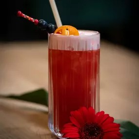 Fruchtiger Cocktail mit Orangenscheibe und Cocktailkirsche, serviert im hohen Glas mit Strohhalm – dekoriert mit einer roten Blüte.

Details: