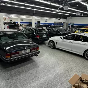 A well-lit garage or showroom displays several luxury vehicles, including a classic black Rolls-Royce in the foreground and various other cars.