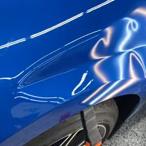 A noticeable dent is visible on the bright blue fender of a car, reflecting light and showing the distorted body line.
