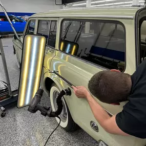 A technician uses specialized tools and a light board to perform paintless dent repair on the rear quarter panel of a light-colored vintage station wagon.