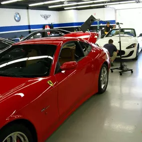 A red Ferrari 612 Scaglietti is parked in a detailing shop with other luxury cars, including a white Maserati, undergoing work by technicians.