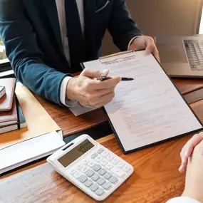 A legal advisor from Ibrahim & Dobaria Law, PLLC discusses a property agreement with a client. The desk displays legal documents, a model house, calculator, laptop, and a notary stamp.