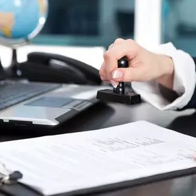 A legal professional at Ibrahim & Dobaria Law, PLLC is stamping an official document on a clipboard, signifying authentication or approval, with a laptop and globe in the background.