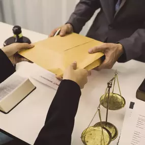 Two professionals exchange a legal envelope across a table with contract documents and scales, symbolizing Ibrahim & Dobaria Law, PLLC’s dedication to secure transactions, legal agreements, and trusted representation.