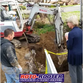 Roto-Rooter crew members are using an excavator to dig near a residential property while discussing the project with a homeowner, likely for a major plumbing or sewer line repair.