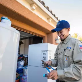 A smiling technician from Lee’s Air, Plumbing, & Heating prepares tools next to a water heater in a garage, wearing company uniform and cap.