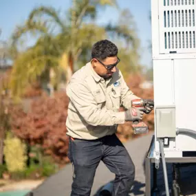 A Lee’s Air technician uses a power drill to service an HVAC unit on a rooftop, surrounded by palm trees and blue sky.