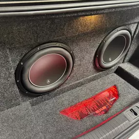 The trunk of a car is customized with two large car audio subwoofers, expertly installed in a gray carpeted enclosure. A red reflective safety triangle is visible on the floor between them.