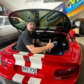 A man wearing a cap and glasses is smiling and giving a thumbs-up from the open trunk of a red sports car with white racing stripes. The car is in a garage or workshop setting.