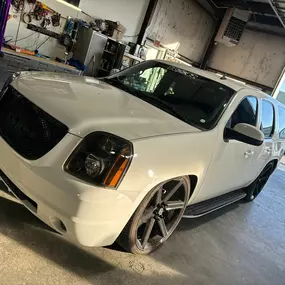 A white customized SUV with dark wheels is parked on a polished concrete floor in a garage or workshop. Tools and equipment are visible in the background, suggesting a vehicle customization business.