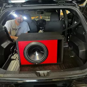 The trunk of a black SUV is open, revealing a large, red, rectangular subwoofer enclosure with a black speaker. A person in a grey shirt is visible in the background, working near the vehicle.