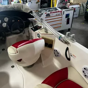 The interior of a white boat features white and red cushioned seats and a gray steering wheel with gauges. Speakers are visible on the boat's side, and a toolbox and other equipment are in the blurred background.