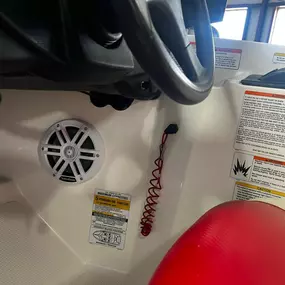 The interior of a boat's helm station shows a white speaker, a red coiled safety lanyard, and various warning labels. A gray steering wheel and ignition key are also visible, suggesting a close-up of the cockpit.