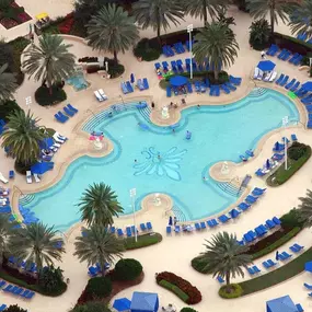 Aerial view of a resort-style pool with a curvy design surrounded by blue lounge chairs and umbrellas. Palm trees add a tropical feel.