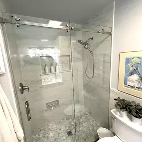A modern bathroom shower stall with a frameless, stainless steel sliding glass door. The walls and floor are tiled in a light gray/white marble-look tile.