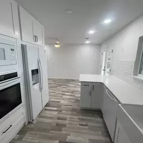 A bright, modern kitchen features white cabinetry, appliances, and countertops. The flooring is a light gray and beige wood-look tile.