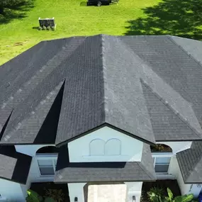 Aerial view of a house with a dark gray, multi-gabled roof. The yard is green and features a trailer and shrubs, conveying a neat, suburban setting.