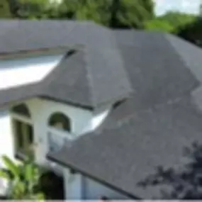 High-angle close-up view of a house. The house’s roof is covered with dark gray asphalt shingles. The roof has complex shapes and angles.