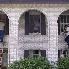Three people are cleaning second-story windows of a white brick house using ladders.