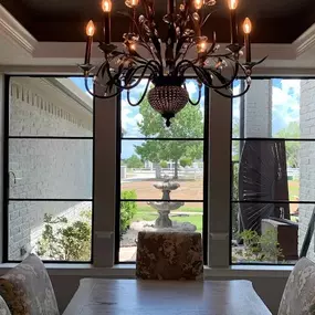A dining area with large windows featuring black metal frames. The aluminum windows are positioned in a corner configuration, offering expansive views of a landscaped yard and rural scene beyond.