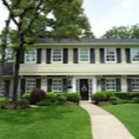 A large, light beige-colored house with a dark gray roof is situated on a grassy lawn. The house features numerous evenly spaced, white vinyl windows with matching dark brown/black shutters.