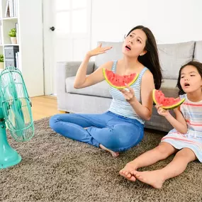 A mother and daughter sit together in their warm home, using a fan in an attempt to stay cool during hot weather. The daughter holds her hair back while the fan blows air in their direction, and both show visible signs of discomfort from the heat—suggesting the air conditioning may not be working properly. This image highlights the need for reliable A/C systems, fast HVAC repair, and energy-efficient cooling solutions to maintain comfort and safety indoors.
