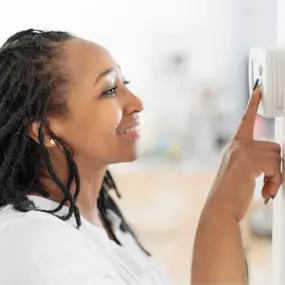 A woman stands near a wall-mounted digital thermostat, adjusting the temperature settings in her home for optimal comfort. Her focused expression suggests she’s trying to cool the house down or warm it up, depending on the season. This image illustrates everyday HVAC use, energy-saving habits, and the importance of a responsive heating and cooling system. Perfect for promoting thermostat upgrades, smart HVAC controls, or seasonal maintenance services.