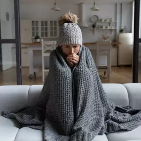 A woman bundled up in a hat and jacket, sits in her chilly home trying to stay warm. She appears uncomfortable, layered in winter clothes, and clearly affected by the cold indoor temperature—possibly due to a broken heater or inefficient furnace. This image perfectly illustrates the importance of reliable home heating, timely HVAC maintenance, and professional furnace repair during the winter months. Great for use in heating service promotions or cold weather safety tips.
