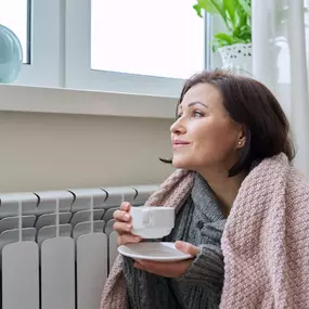A woman wrapped in a cozy blanket sits indoors, holding a steaming cup of coffee or tea, trying to stay warm during cold weather. Dressed in layered clothing and thick socks, she’s clearly feeling the chill—possibly due to a broken heater or an inefficient heating system. The scene highlights the importance of reliable home heating and indoor comfort during winter. Ideal visual for HVAC repair services, furnace maintenance, heating system installations, or energy-efficient heating solutions.